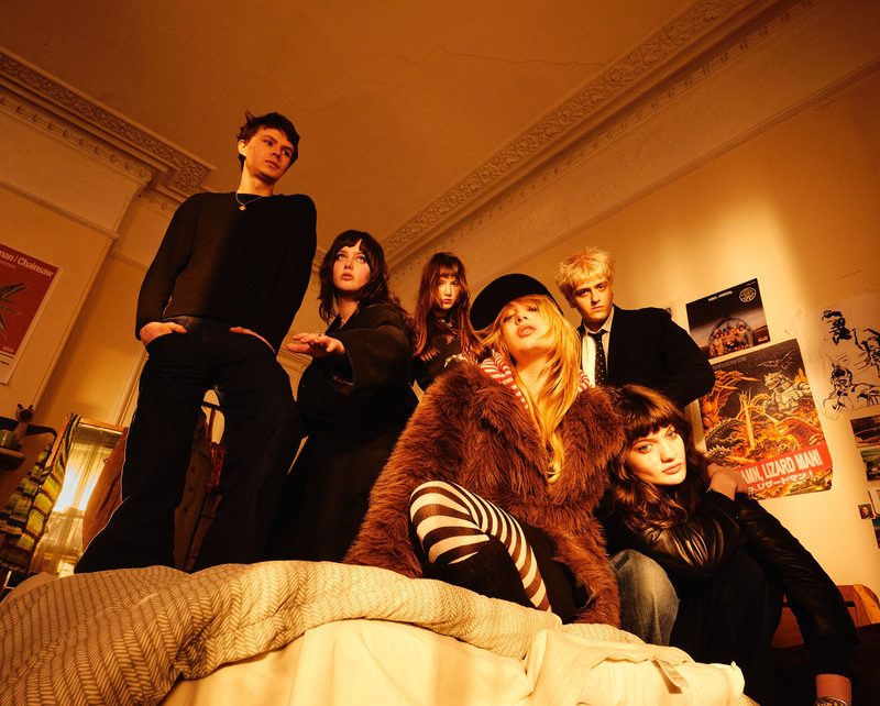 The six members of Man/Woman/Chainsaw pose in a room lit in orange tones