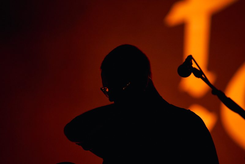 The silhouette of a Fontaines D.C. member appears beside the microphone against orange light in Alicante, Spain.