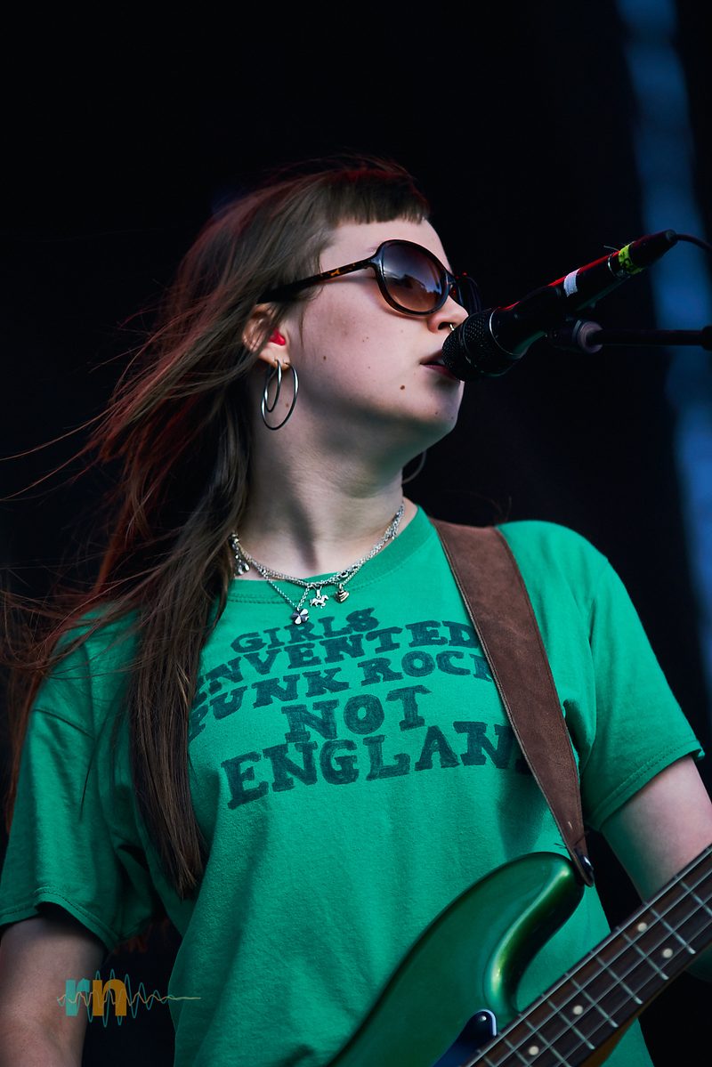 La bajista de Man/Woman/Chainsaw canta al micrófono con camiseta verde y gafas de sol en Canela Party 2026.
