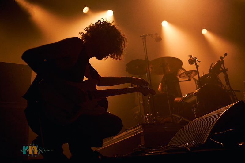 The MAQUINA. guitarist plays crouched with the drummer behind him under amber light at Canela Party 2026.